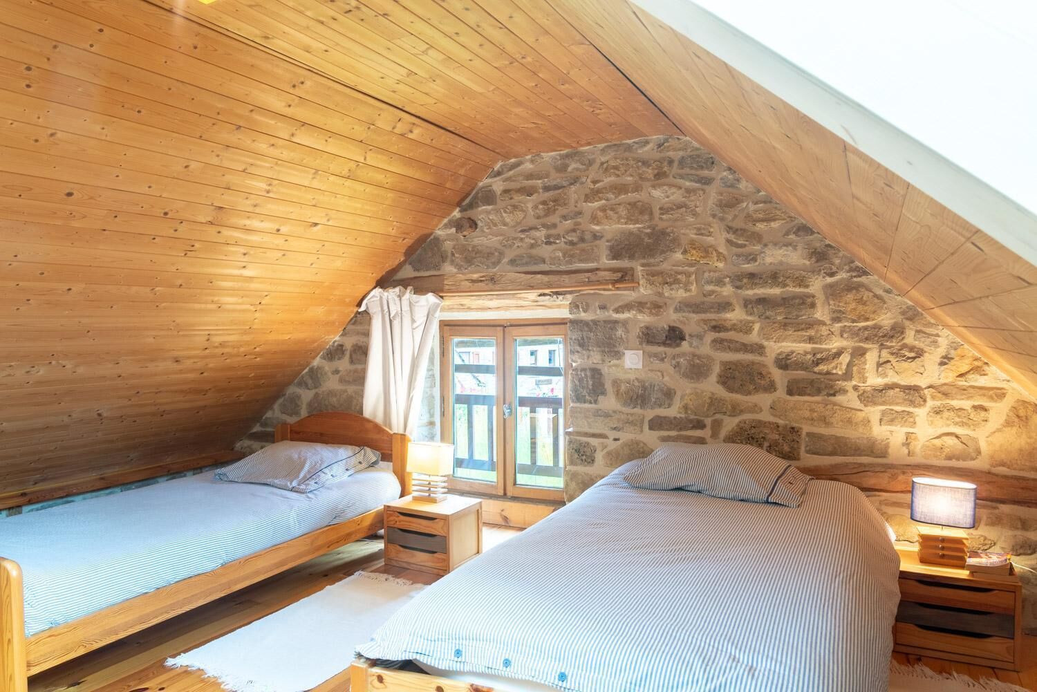 A cozy attic bedroom with two wooden beds, stone walls, and a window with curtains.