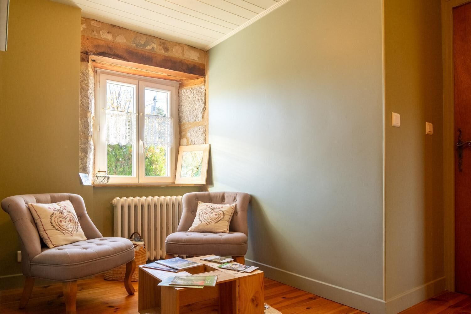 Ein Zimmer mit zwei Sesseln, einem Holztisch, einem Heizkörper und einem Fenster mit Spitzenvorhängen.