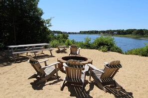 Ein Sandstrandabschnitt mit Feuerstelle, Holz-Adirondack-Stühlen und einem Picknicktisch.