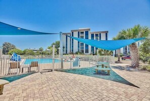 A swimming pool area with a fence, a shaded area, and a building in the background. - Enclave 701a- Shore Fun (Destin)