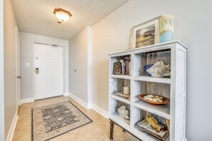 A hallway with a white door, a wooden bookshelf with decorative items, and a patterned rug. - Enclave 701a- Shore Fun (Destin)