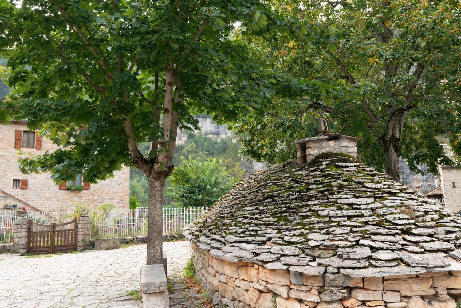 A stone-roofed building with a stone wall, surrounded by trees and a stone fence.