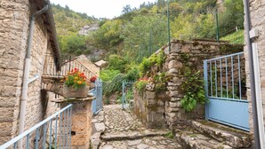 A narrow stone pathway leading to a stone building with a metal gate, surrounded by greenery and a mountainous backdrop. - Gîtes de France 3/5 pers in Saint-Chély-du-Tarn (SAINT-CHELY-DU-TARN)