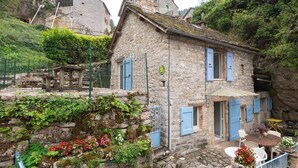 A stone building with blue shutters, a small outdoor seating area, and a stone wall with plants. - Gîtes de France 3/5 pers in Saint-Chély-du-Tarn (SAINT-CHELY-DU-TARN)