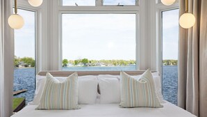Una cama con sábanas blancas y almohadas a rayas, un ventanal con vistas a un lago y casas, y lámparas colgantes.