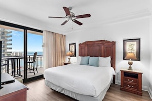 A hotel room with a large bed, a ceiling fan, a balcony with a view of the ocean, and a desk with a chair. - Emerald Grande W522- Harbor Breeze (Destin)