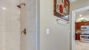 Ein Badezimmer mit Dusche, Toilette und Waschbecken. An der Wand hängt ein Bild und durch eine offene Tür ist eine Küchenzeile erkennbar.