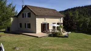 A two-story house with a patio, a dining area with outdoor furniture, and a forested hillside in the background. - The Parrot in Margeride (PIERREFICHE)