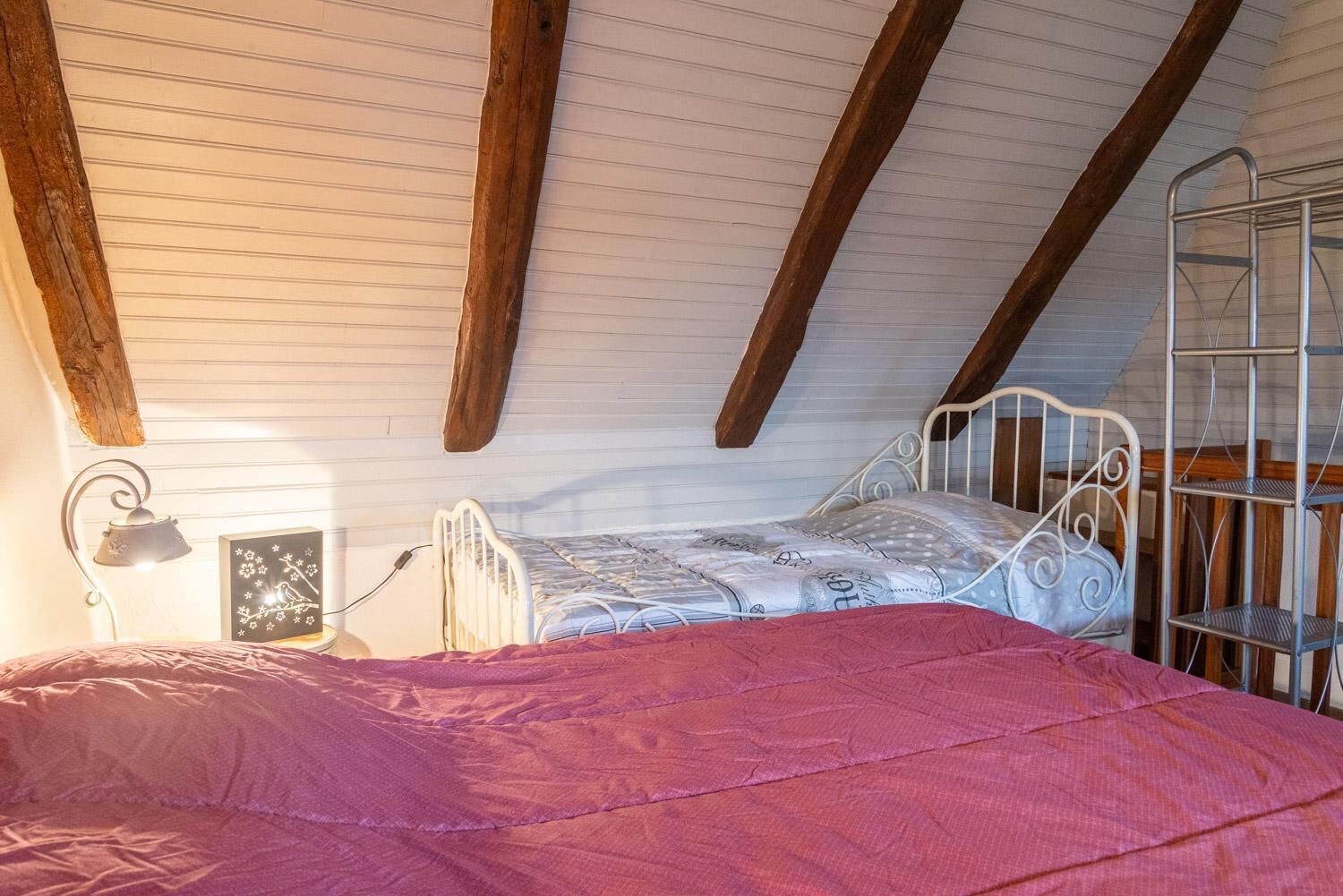 A bedroom with a sloped ceiling, a metal bed frame, a bedside lamp, and a metal shelf unit.