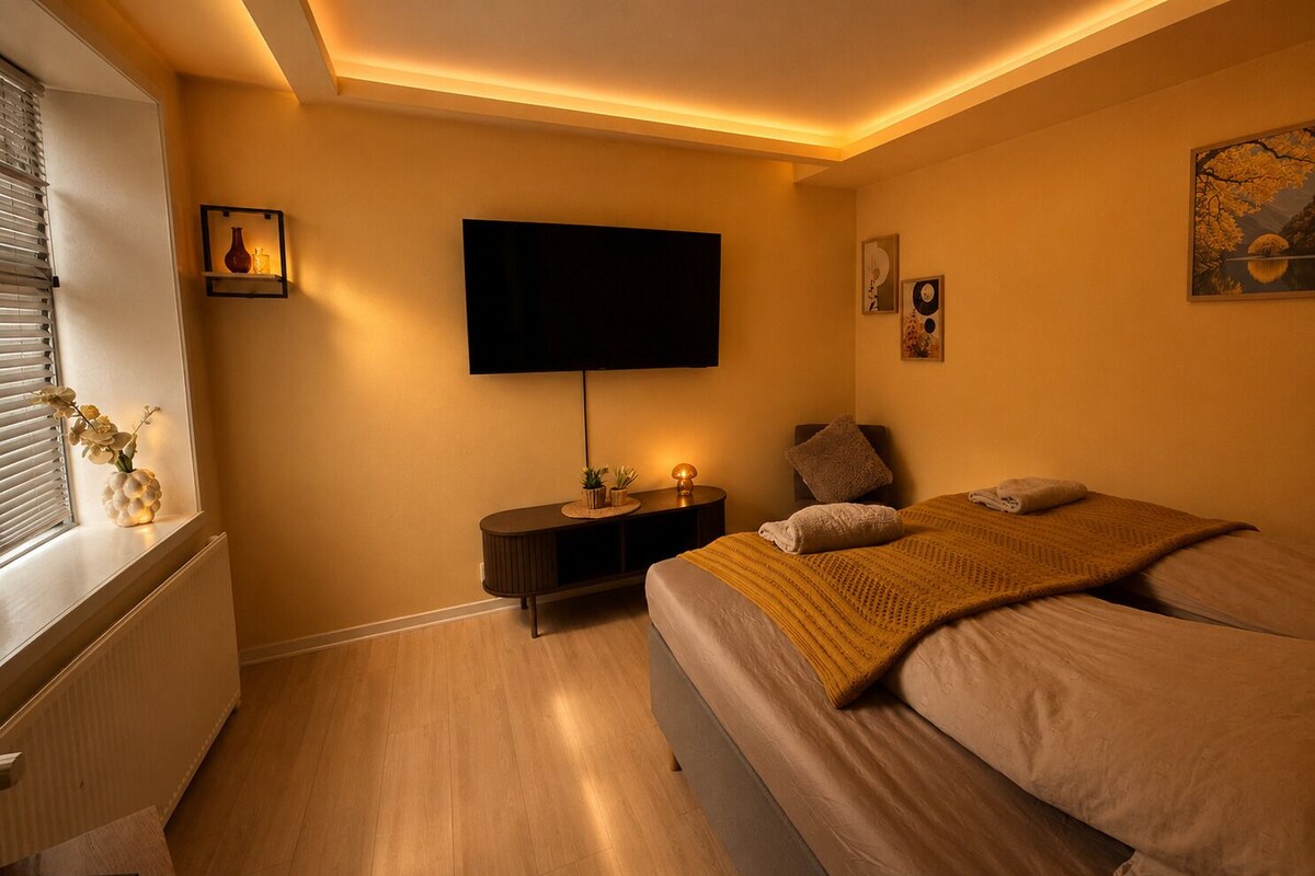 Un dormitorio con una cama, un televisor montado en la pared, una mesita de noche con una lámpara y una ventana con persianas.