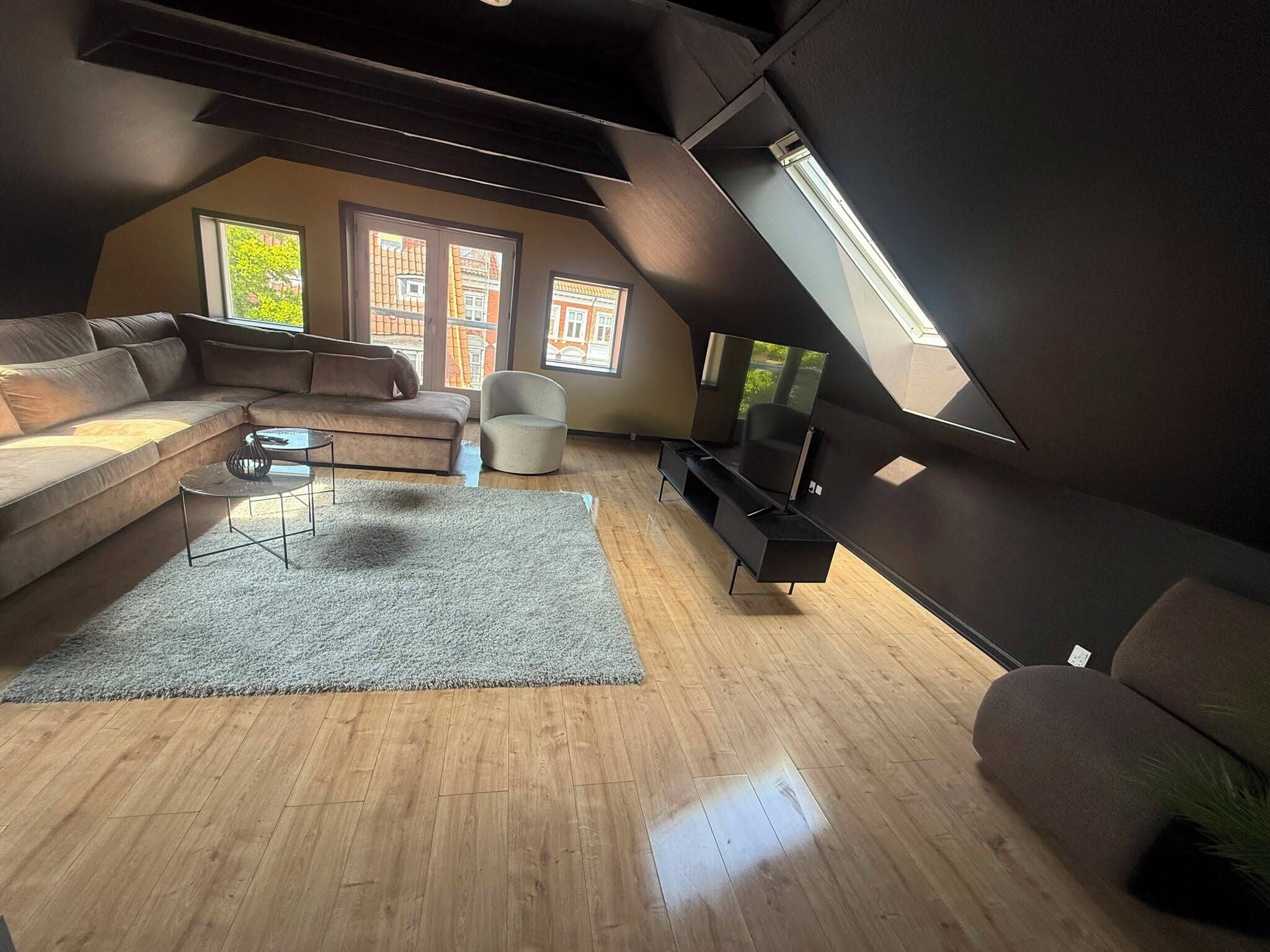 A modern loft-style living room with a sloped ceiling, a large sofa, a coffee table, a TV stand, and a small chair.