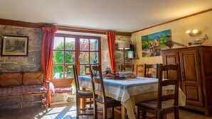 A dining area with a table set for two, wooden chairs, a stone wall, a window with curtains, and a painting on the wall. - Gîte La Narce - TERMES (TERMES)