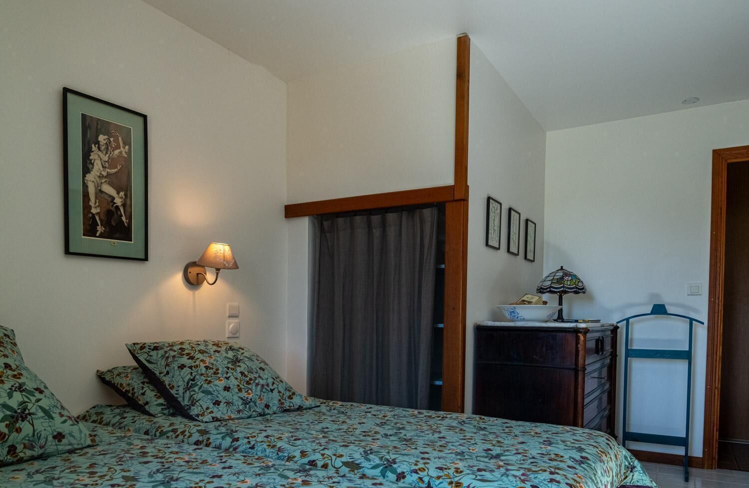 Een slaapkamer met een bed, nachtkastje, lamp, een lijst met een foto aan de muur en een deur.