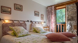 A bedroom with two beds, floral bedding, framed pictures on the wall, a window with curtains, and a stone wall. - Gîte La Narce - TERMES (TERMES)