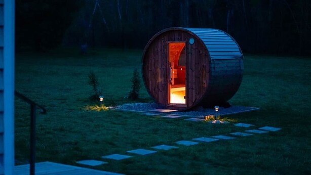 Una estructura de madera con forma de barril, con una puerta abierta, un pequeño espacio con asientos al aire libre y un sendero que lleva a ella.