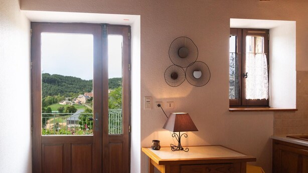 Una habitación con puerta de madera, ventana con cortinas de encaje, lámpara fijada a la pared y vistas a un paisaje verde.