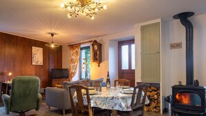 Un salon chaleureux avec une cheminée, une table à manger dressée pour deux personnes, un fauteuil vert et un téléviseur.