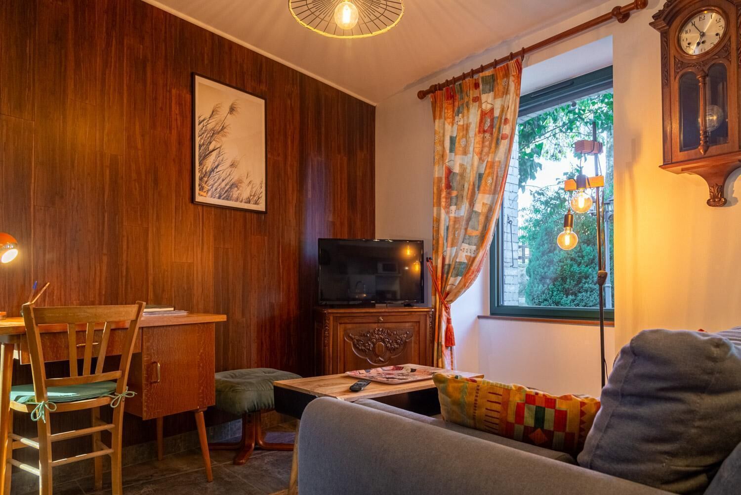 A cozy room with a wooden wall, a television, a sofa, a table, and a window with curtains.