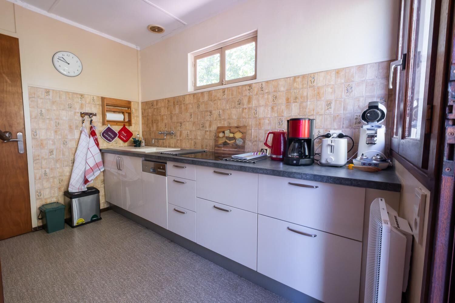 A compact kitchen with a sink, stove, and coffee maker. There is a clock on the wall and a window above the sink.