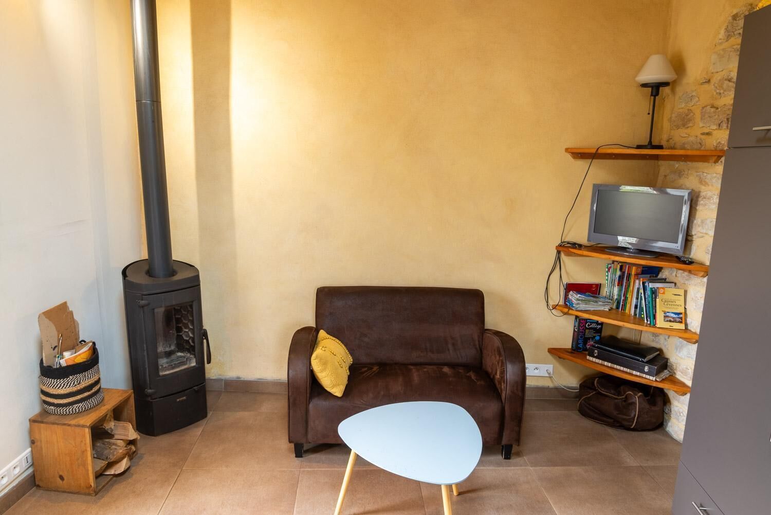 Una sala de estar con un sofá de cuero marrón, una mesita blanca pequeña, un televisor sobre una estantería de madera y un estufa de leña.