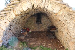 A stone-arched structure with a hearth, scattered firewood, and a metal rack. - Gîte du Pigeonnier Causse Méjean (SAINT-PIERRE-DES-TRIPIERS)