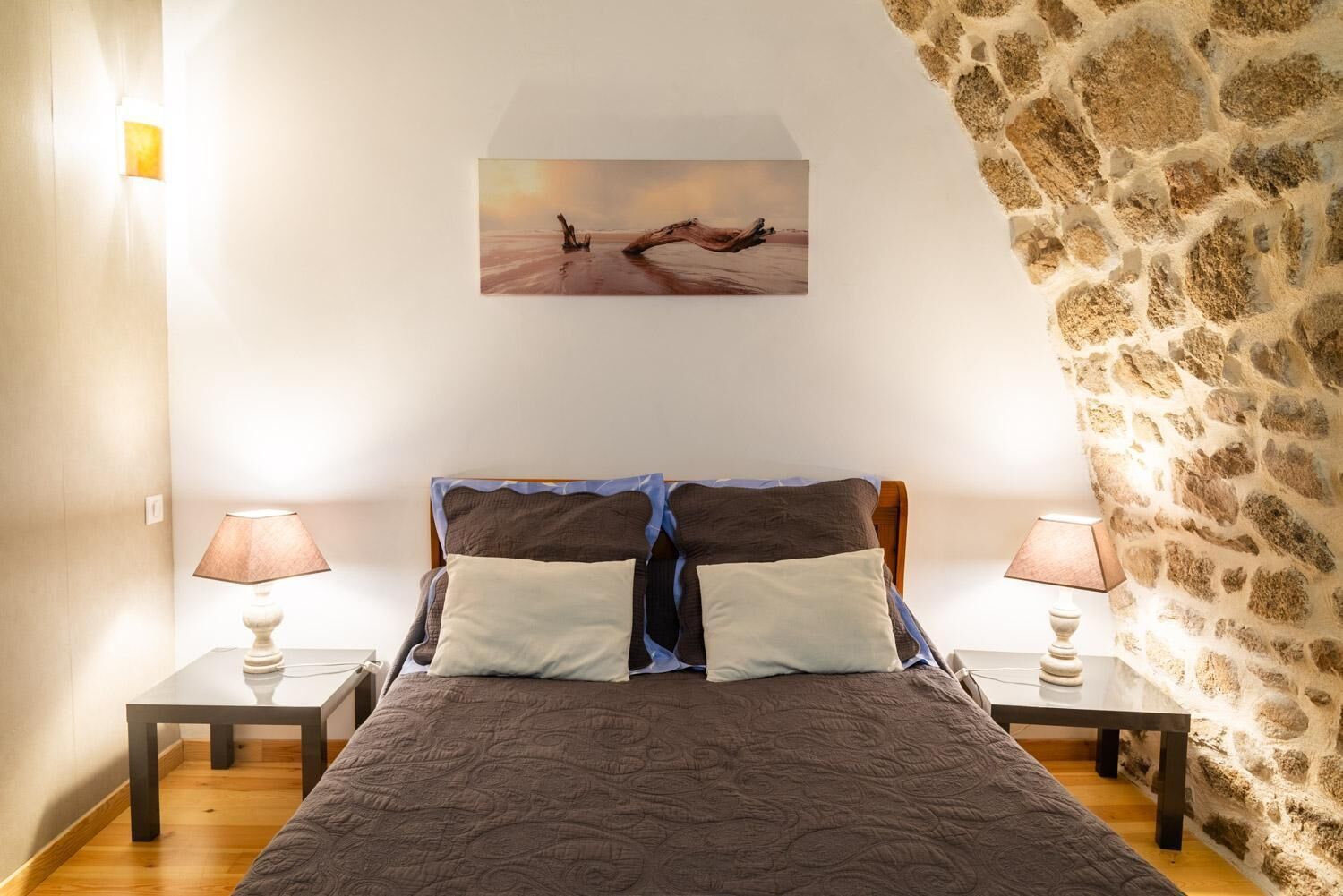 A bedroom with a bed, two bedside tables, a lamp, a painting, and a stone wall.