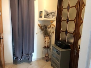 A room with a wooden door, a grey curtain, a vase with dried flowers, a small table with a teddy bear, and a cabinet. - La Glycine 5/6 persons (LA CANOURGUE)