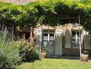A garden with a pergola, potted plants, and a building with a door and windows. - La Glycine 5/6 persons (LA CANOURGUE)