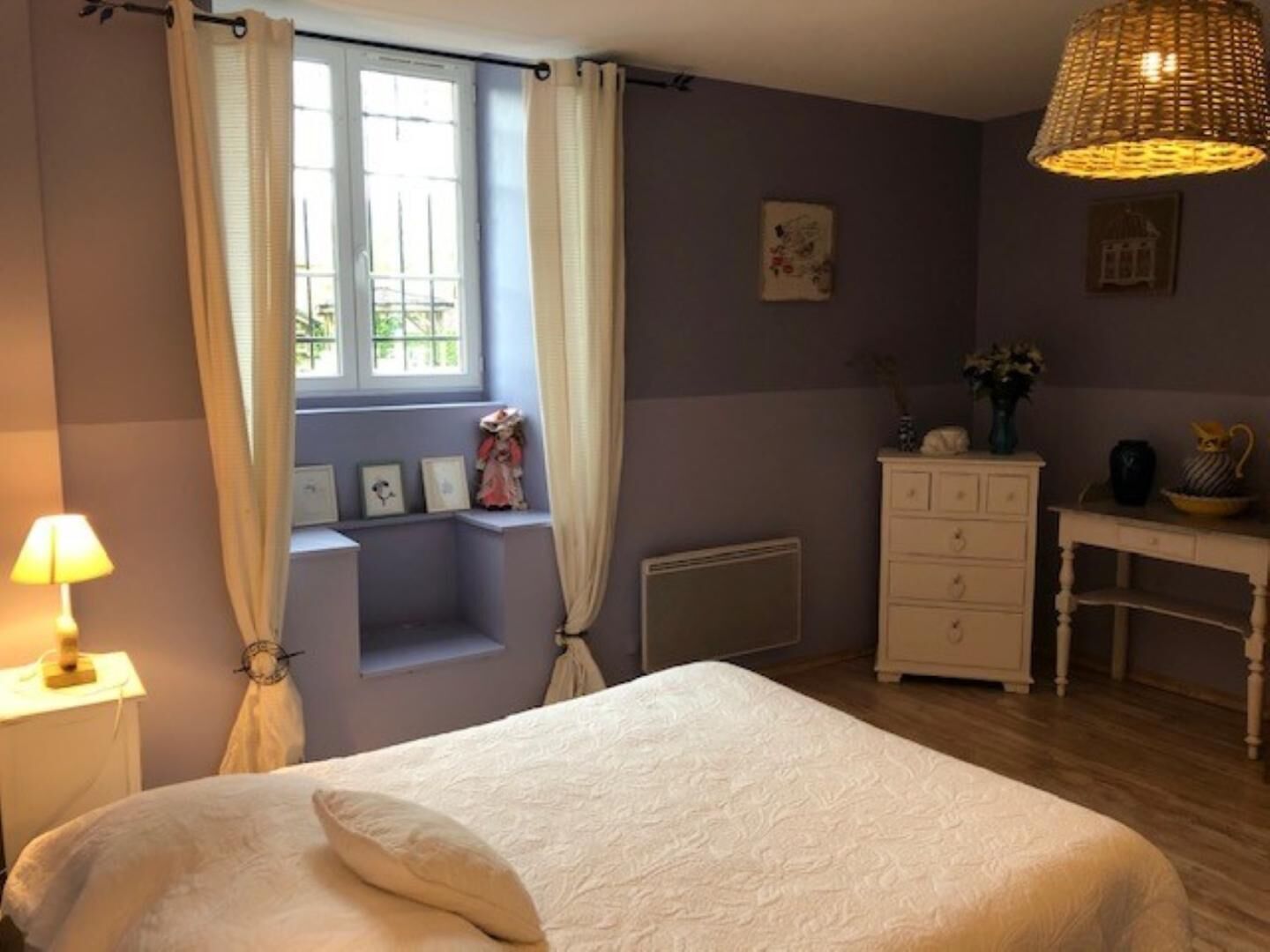 Un dormitorio con cama, cómoda, escritorio, ventana con cortinas y lámpara colgante.