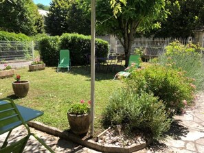 A garden area with a lawn, potted plants, a metal table, and a chair. - La Glycine 5/6 persons (LA CANOURGUE)