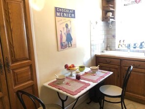 A kitchen with wooden cabinets, a table with a placemat, and a poster on the wall. - La Glycine 5/6 persons (LA CANOURGUE)