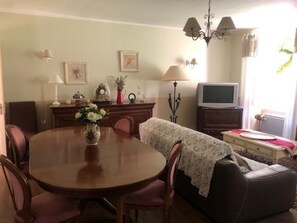 A dining area with a round table, chairs, a vase with flowers, a television, and a sofa. - La Glycine 5/6 persons (LA CANOURGUE)