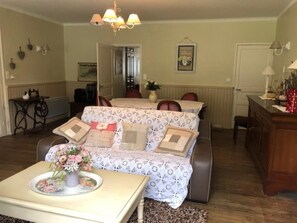 A cozy living room with a sofa, a coffee table with a floral arrangement, and a dining area with a table and chairs. - La Glycine 5/6 persons (LA CANOURGUE)