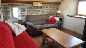 Een gezellige kamer met een stenen muur, een houten plafond, een grijze bank met rode kussens, een houten tafel en een kleine lamp.