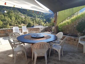 An outdoor patio with a round table and white plastic chairs, surrounded by stone walls and a view of a forested hillside. - La Cazelle - LE ROZIER (LE ROZIER)