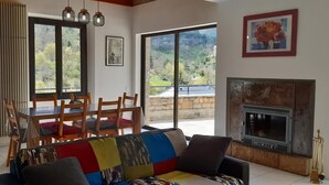 A living room with a sofa, a dining table with chairs, a fireplace, and large windows with a view of greenery. - La Cazelle - LE ROZIER (LE ROZIER)