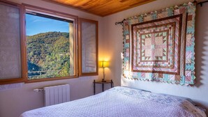 A bedroom with a bed, a window with a view of a forest, a wall hanging, and a bedside table with a lamp. - Les Hauts de Maison Rouge (VIALAS)