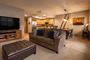 A living room with a brown sofa, a leather ottoman, a flat-screen TV, wooden cabinets, a kitchen in the background, and a dining area with a table and chairs. - Quiet Corner • Walk to Lifts & Main St • Hot Tubs • Private Garage + 1 Reserved (Breckenridge)