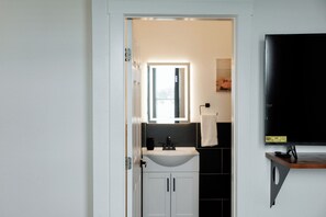Ein Badezimmer mit Waschbecken, Spiegel, Handtuch und einem schwarzen Fernseher auf einem Holzregal.