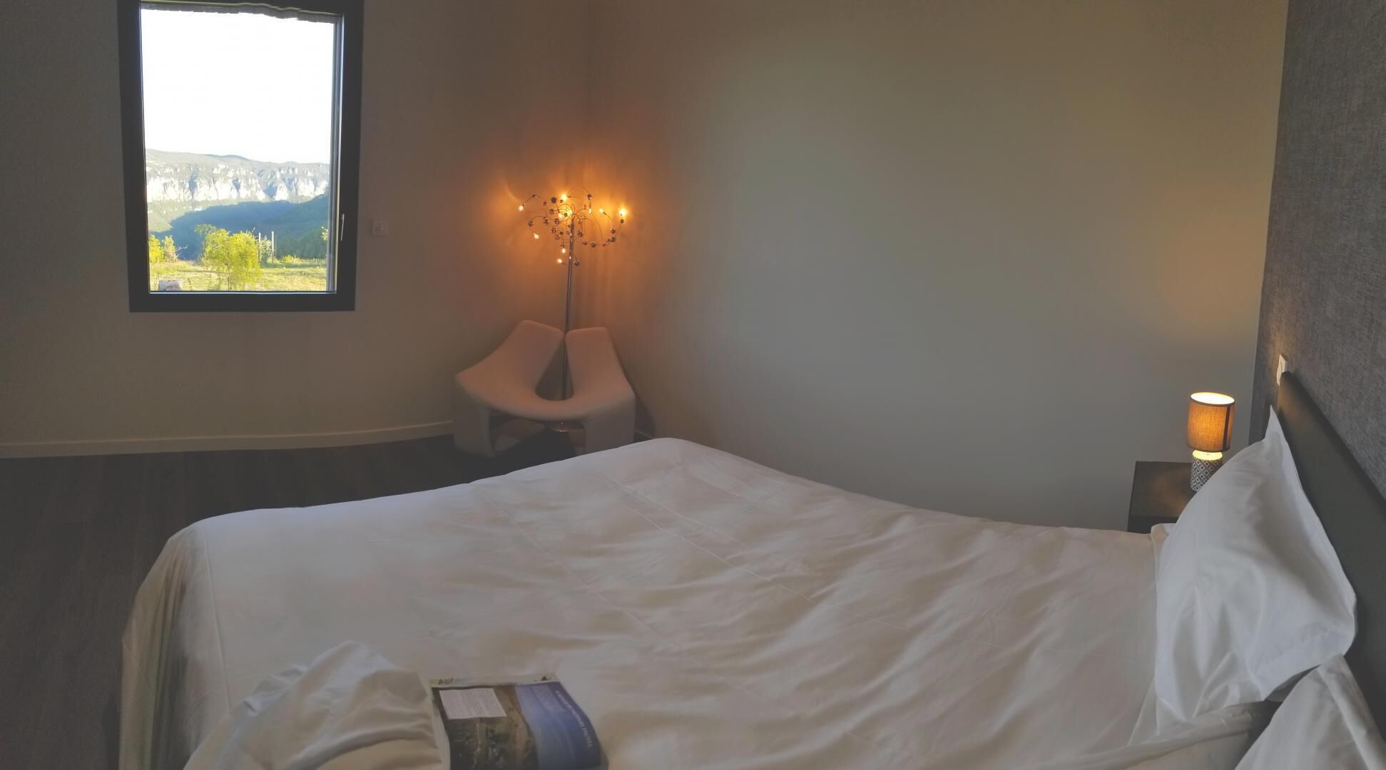 Una camera da letto con un letto, una sedia, una lampada da comodino, una finestra con vista e un punto luce a parete.