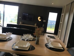 Un comedor moderno con una mesa de madera dispuesta para cuatro, un televisor en la pared y vistas a las montañas por la ventana.