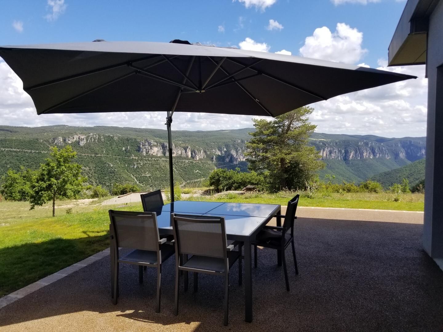 A table with four chairs and a large umbrella, set on a gravel patio with a scenic mountain view.
