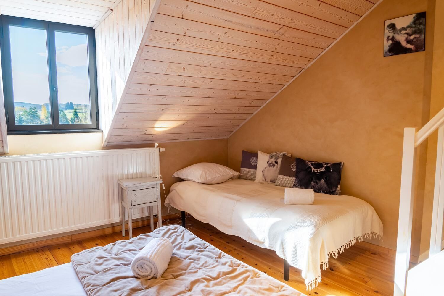 A small, cozy bedroom with a sloped ceiling, a single bed, a bedside table, and a window with a view of greenery.