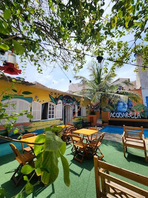 A colorful outdoor seating area with tables and chairs, a mural with the text 'ACQUARELA,' and a pool in the background. - ACQUARELA HOSTEL  (Arraial do Cabo)