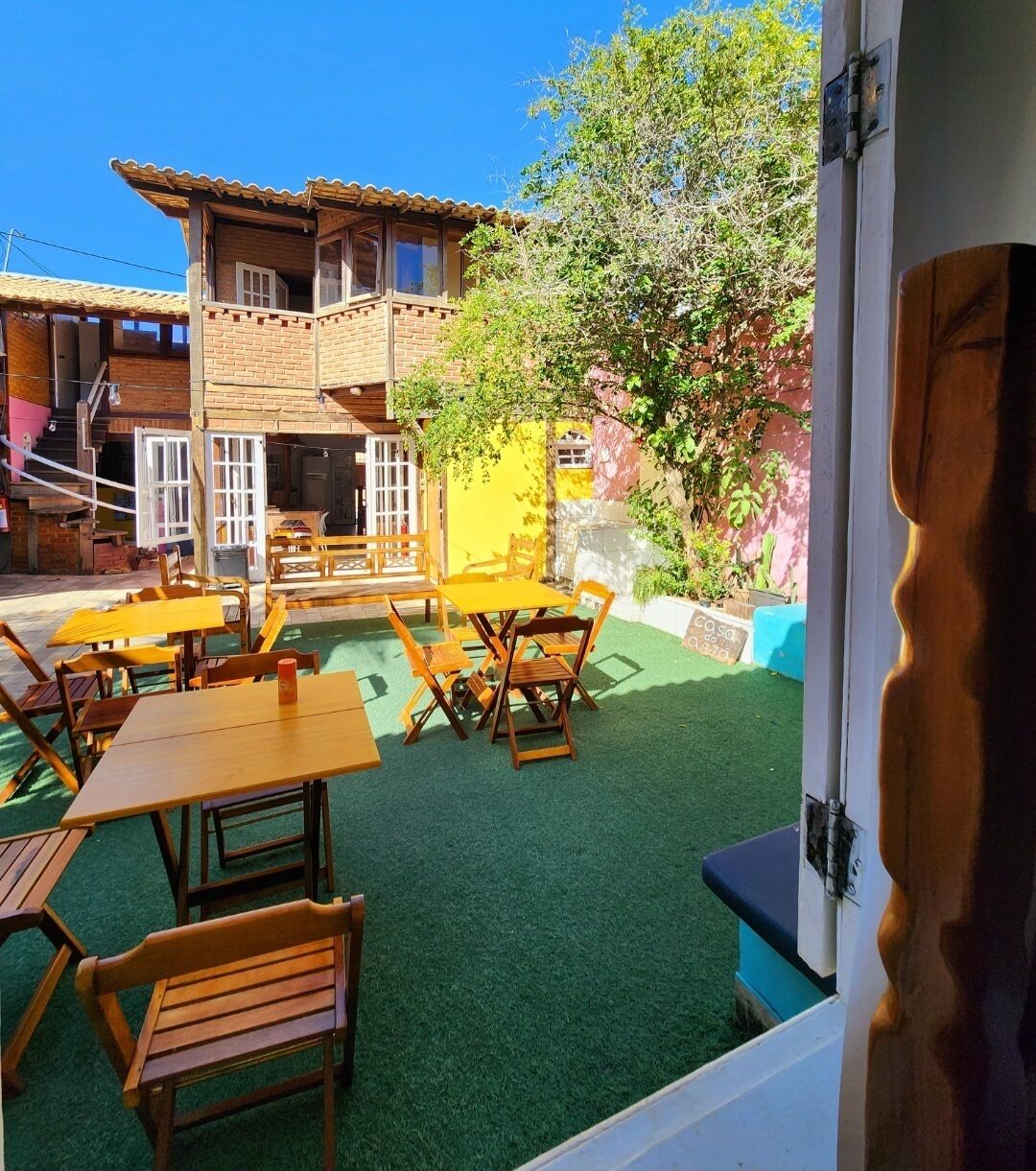 An outdoor patio with wooden tables and benches, a building with a tiled roof, and a green artificial turf area.