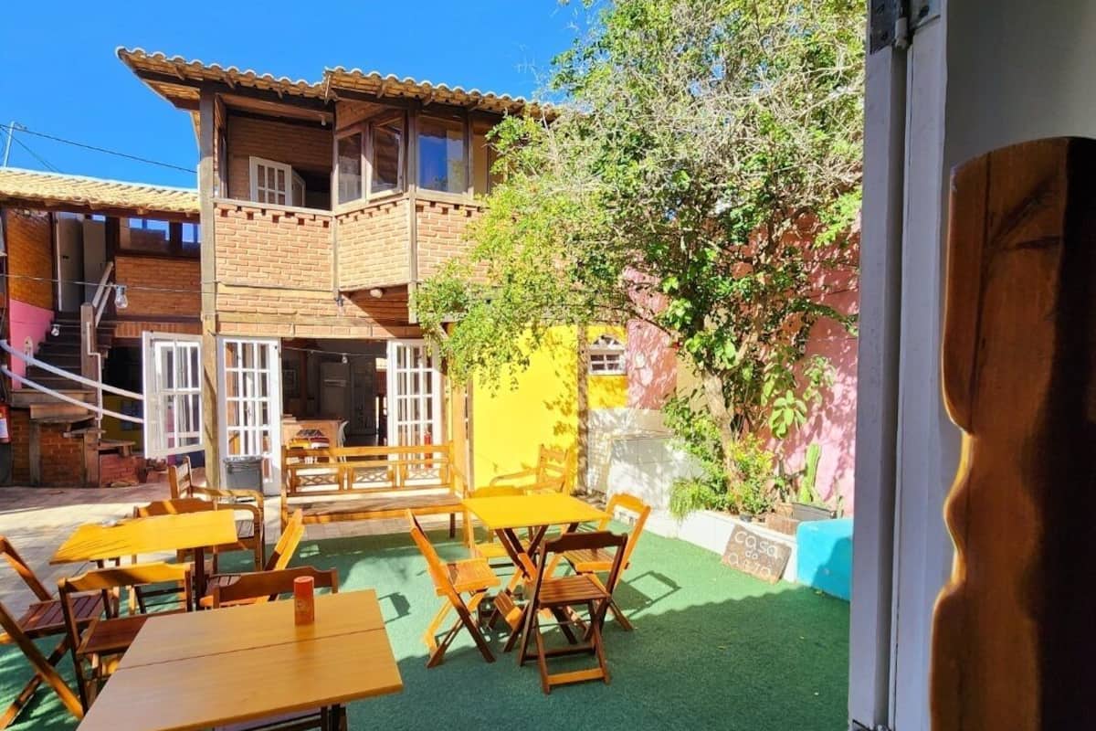 Un patio al aire libre con mesas y bancos de madera, un edificio con tejado de baldosas y una zona de césped artificial verde.