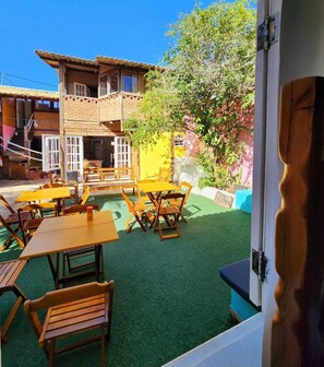 An outdoor patio with wooden tables and benches, a building with a tiled roof, and a green artificial turf area. - ACQUARELA HOSTEL  (Arraial do Cabo)
