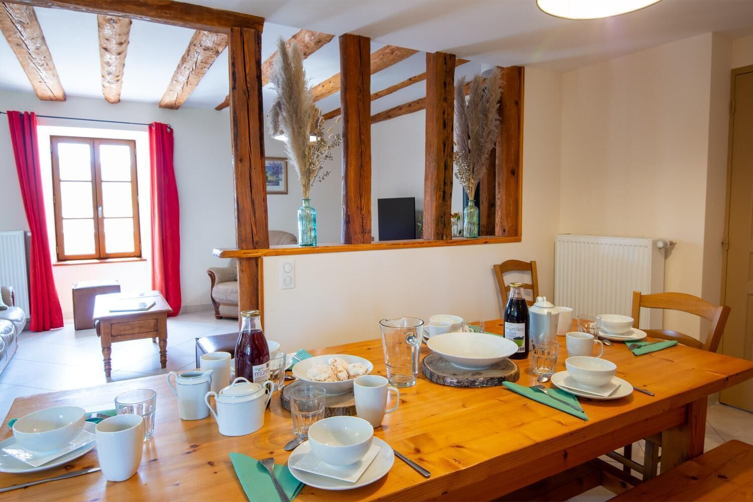 Un coin repas avec une table en bois dressée pour le repas, un miroir au-dessus de la table et une fenêtre ornée de rideaux rouges.