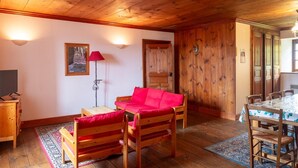 Quarto com painéis de madeira, um sofá vermelho, uma mesa pequena, uma televisão e uma área de jantar com mesa e cadeiras.