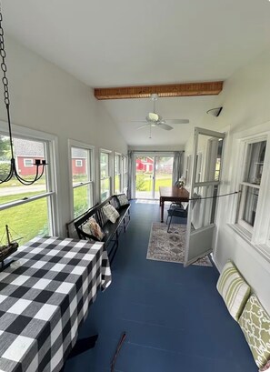 A room with a checkered table, a ceiling fan, and a view of a red house through large windows.